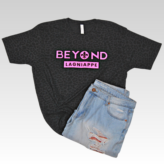 Black Leopard print t-shirt with the Beyond Lagniappe logo across the chest in pink glitter, outlined by black flock vinyl. The "o" in Beyond consists of a filled in letter "o" with a fleur de lis cut out in the center of it. 