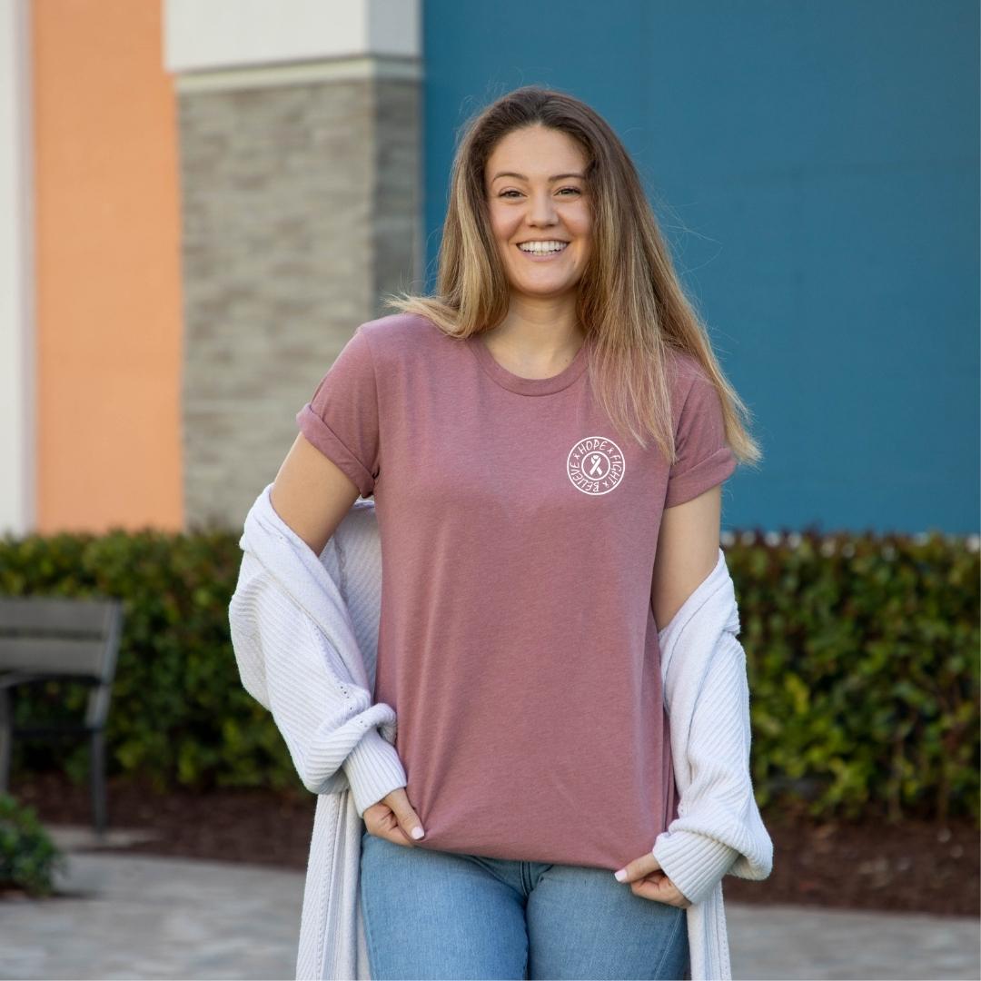 Heather Muave colored cancer support t-shirt front with circle left chest logo in white. The circle is centered with a white ribbon and the words Hope, Fight, Believe going around the inside of the circle. 